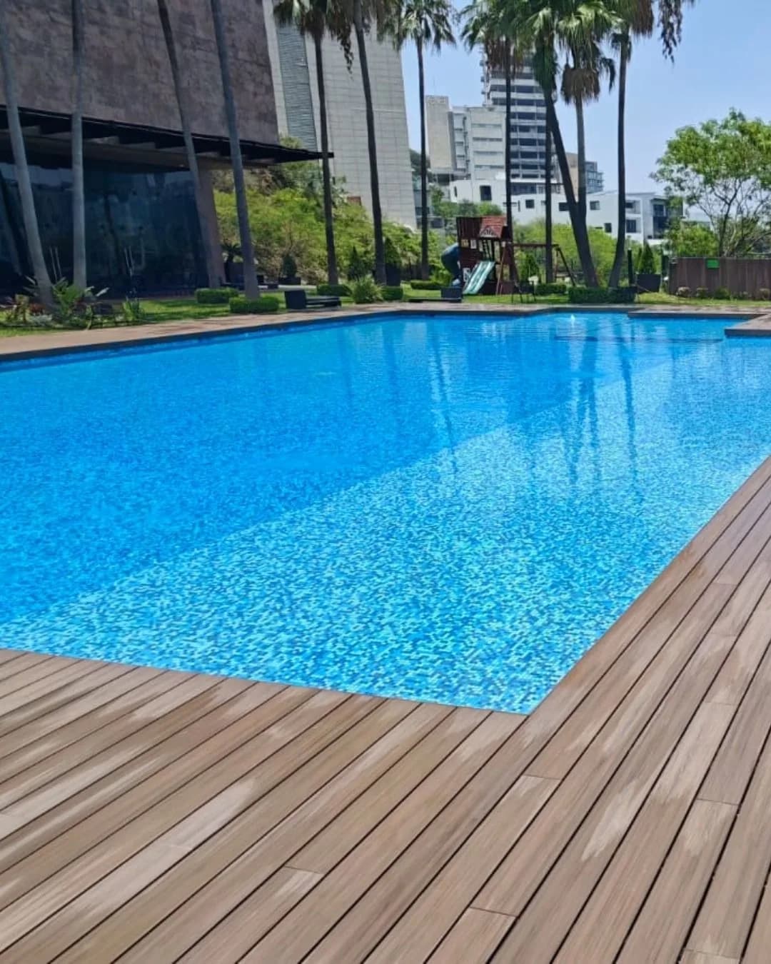Alberca rodeada de deck WPC tono madera junto a un edificio de piedra, con palmeras y juegos infantiles al fondo