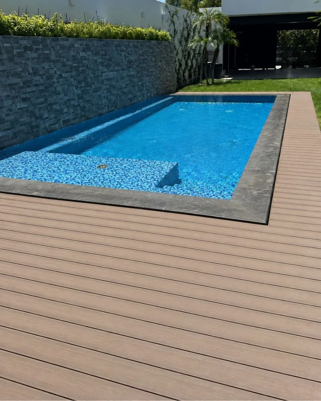 Alberca angosta con deck WPC gris claro a lo largo, junto a un muro de piedra con jardinera