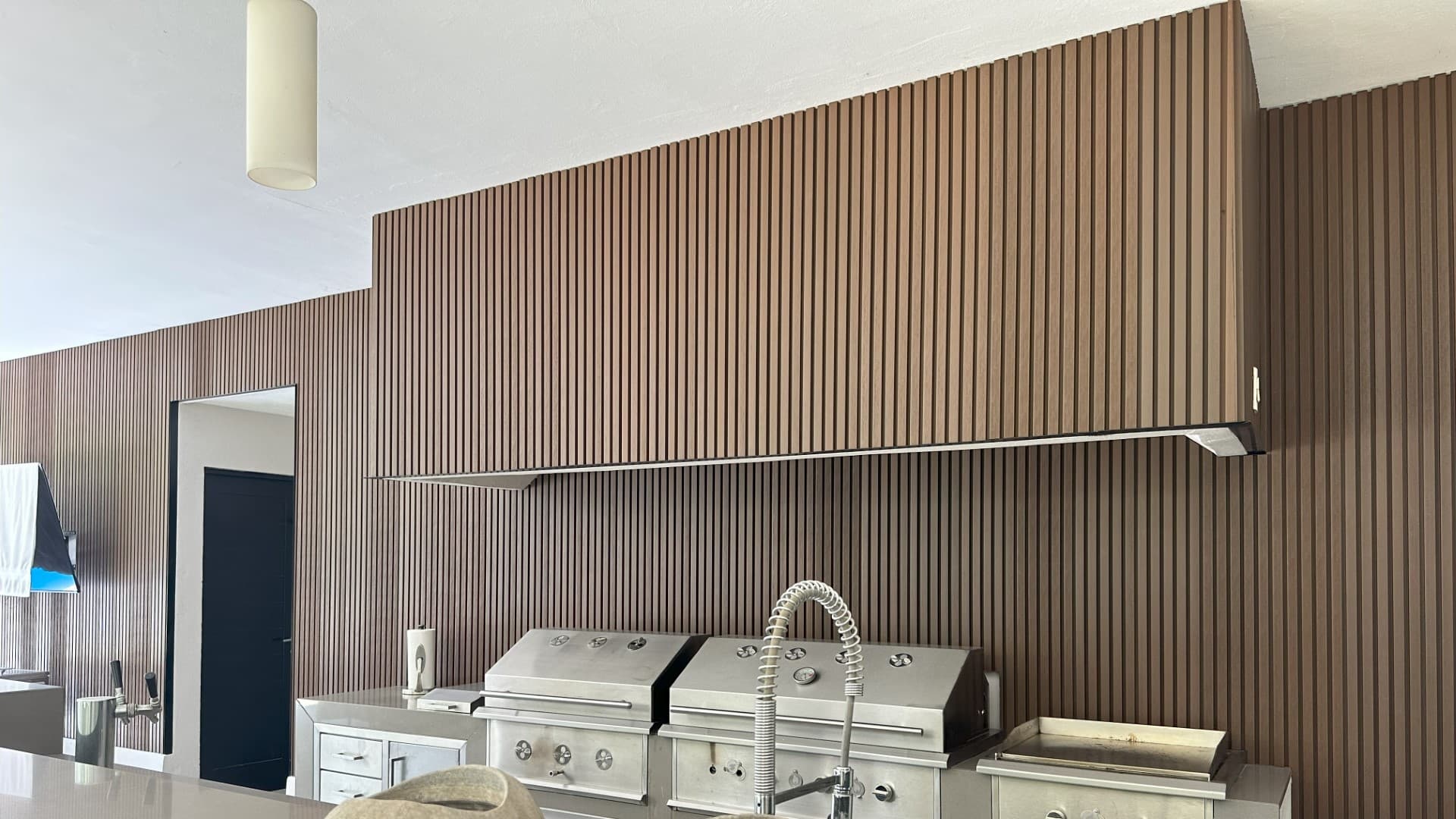 Cocina exterior con asadores de acero inoxidable frente a un muro de lambrín WPC ranurado tono madera