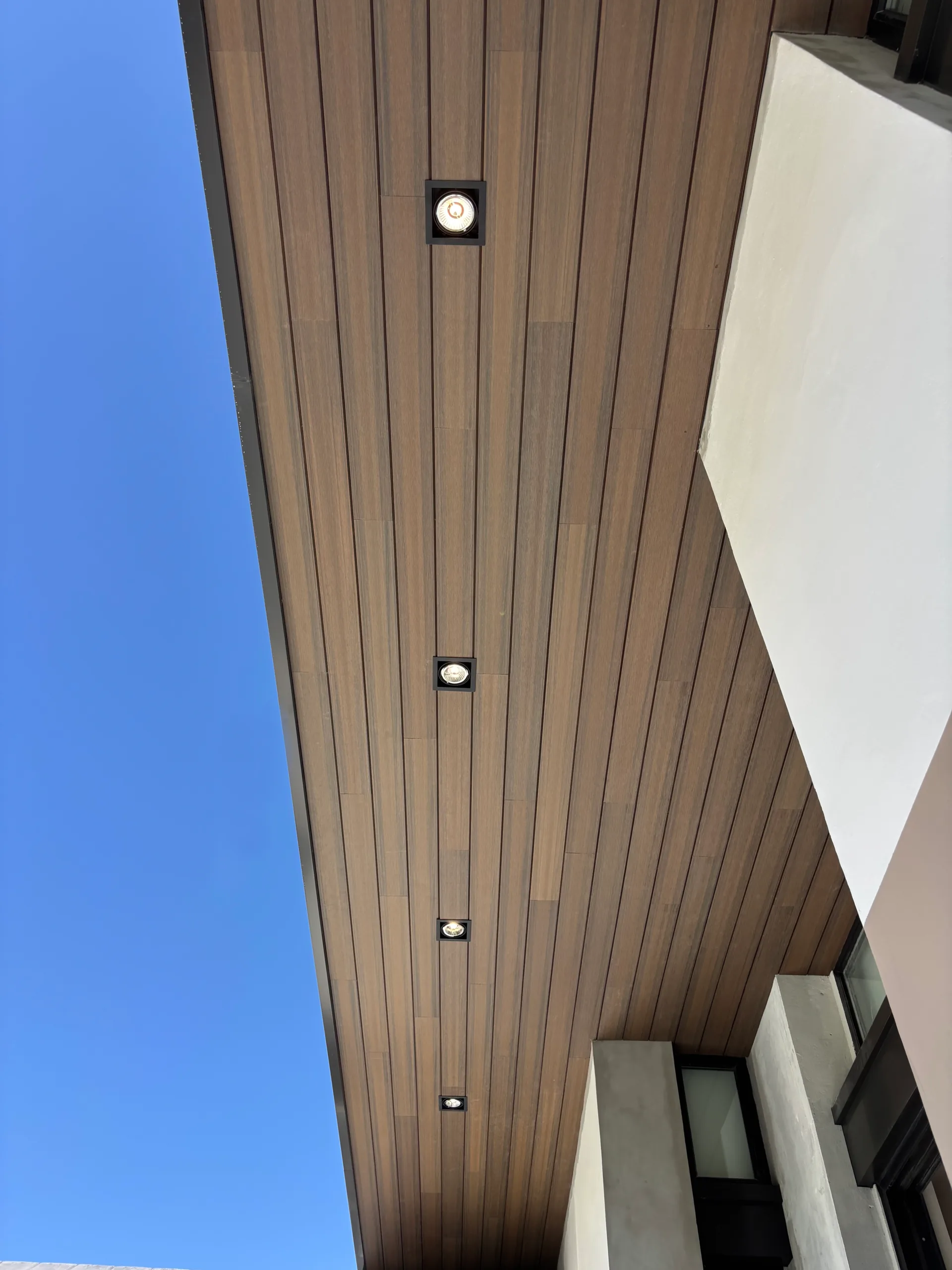 Wall Cladding (Plafón) de WPC tono madera visto desde abajo en el techo exterior de una fachada blanca, con luces empotradas y cielo azul