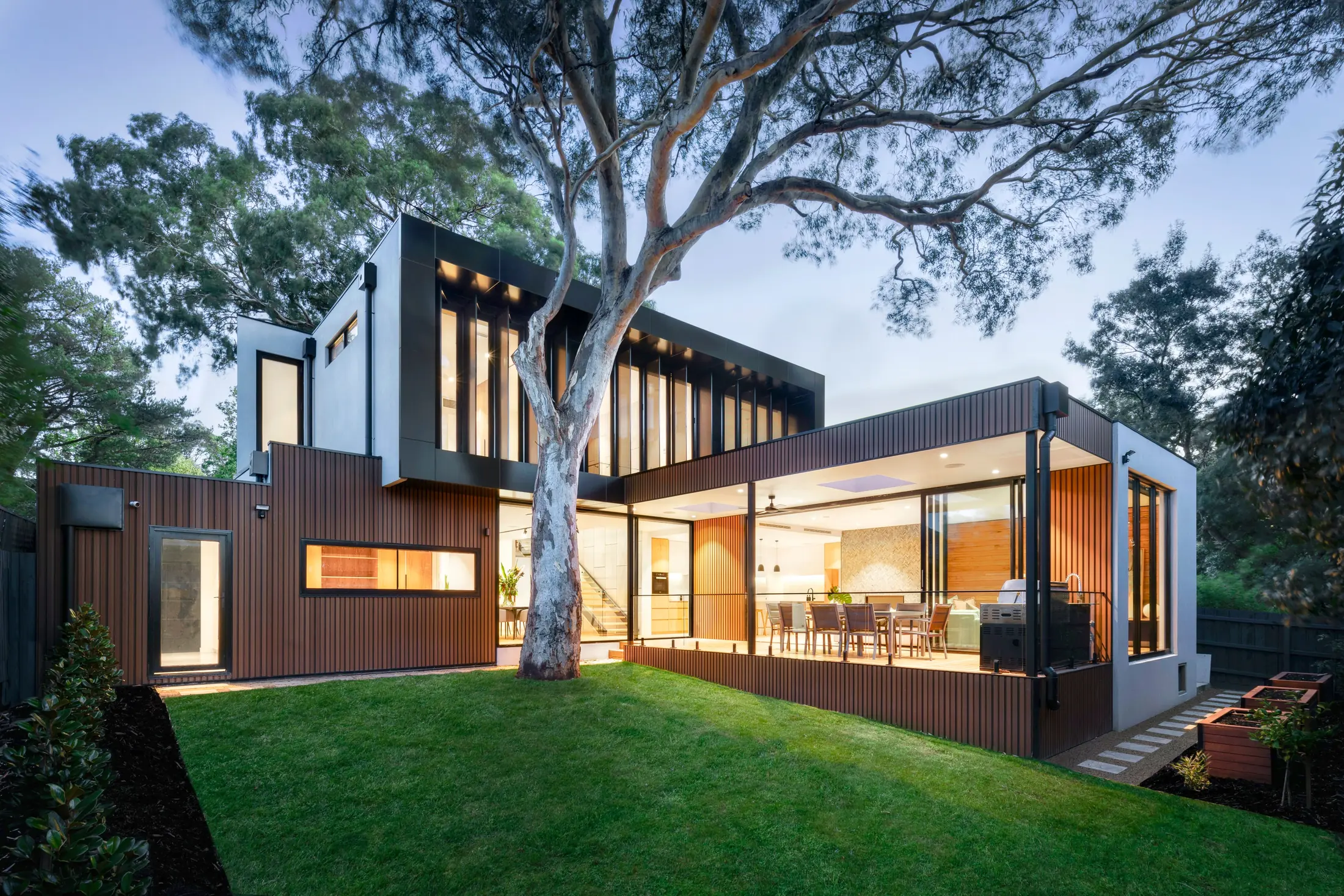 Casa moderna al atardecer con fachada de lambrín WPC tono madera, terraza iluminada y un árbol grande en el jardín