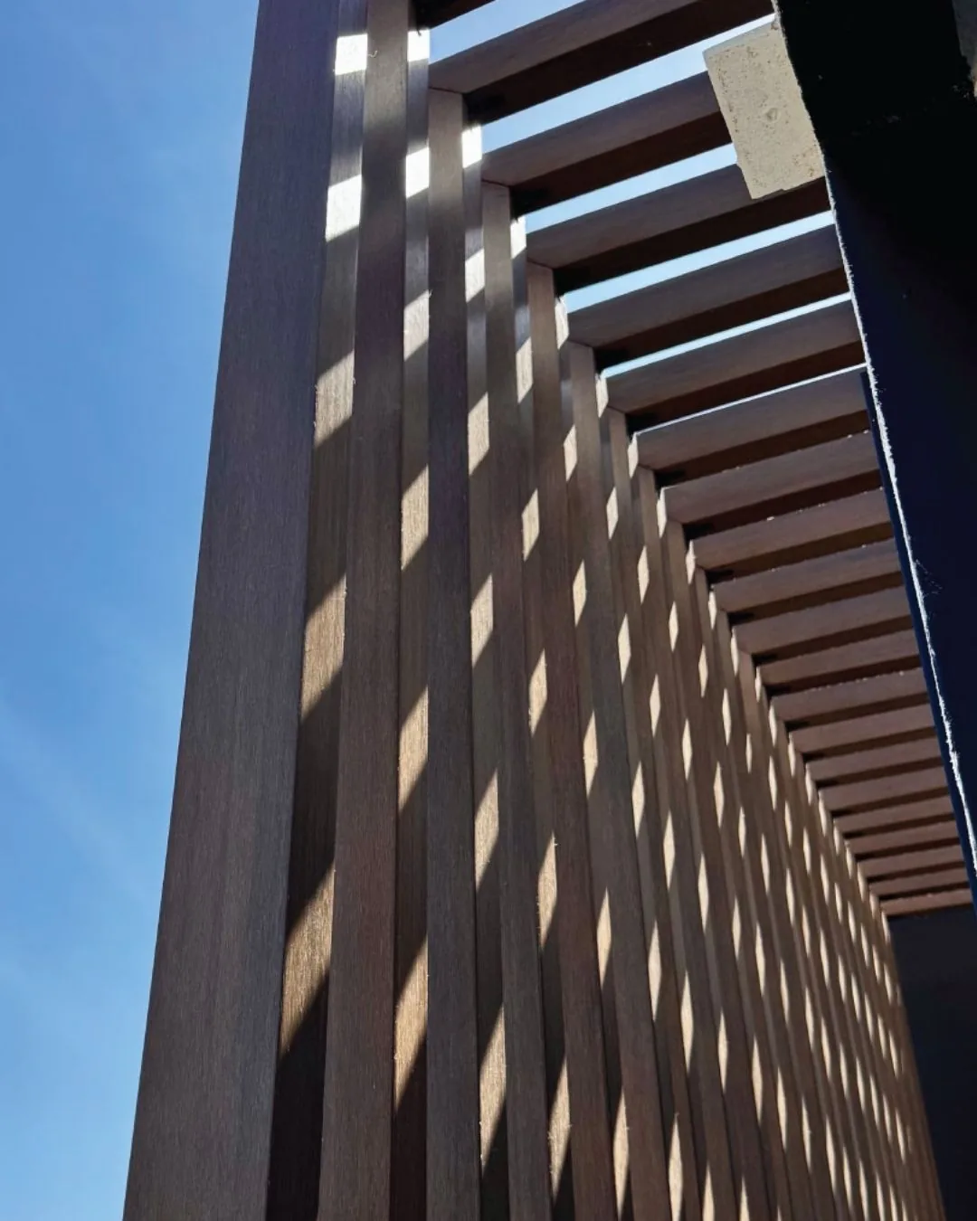 Celosía de vigas decorativas WPC tono madera vista desde abajo contra el cielo azul, con la luz entrando entre las vigas