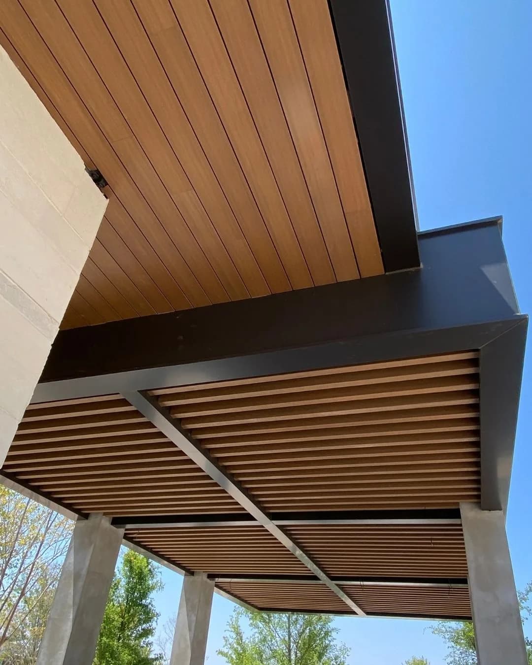 Vigas decorativas WPC tono madera en retícula bajo un volado, vistas desde abajo junto a un plafón de Wall Cladding WPC, con columnas de concreto y cielo azul
