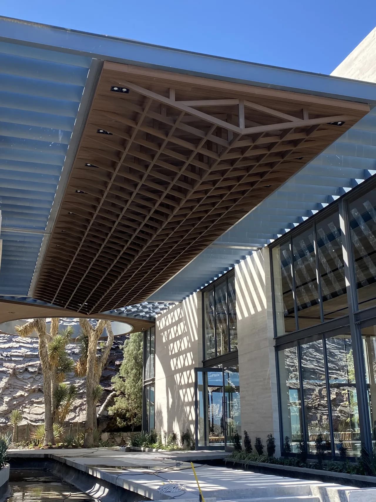 Volado de acceso con una retícula de vigas decorativas WPC tono madera vista desde abajo, junto a una fachada de cristal