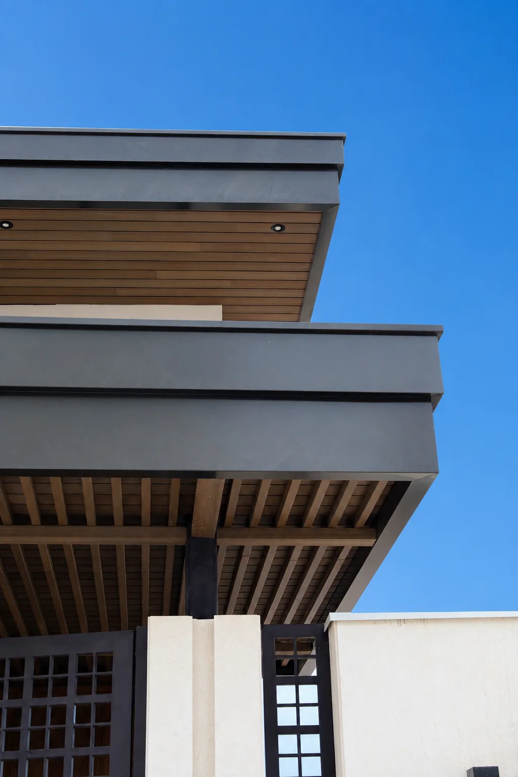Dos volados de una casa vistos desde abajo: el de arriba con plafón de Wall Cladding WPC tono madera y luces empotradas, y el de abajo con vigas decorativas WPC, contra el cielo azul